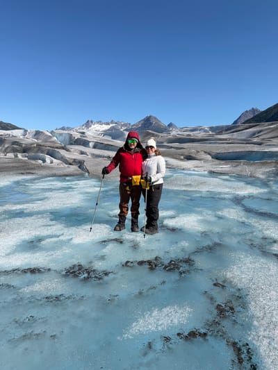 Two people in hiking gear standing on a glacier under a clear blue sky