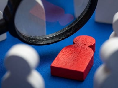 Magnifying glass held over a red wooden figure among white figures on a blue surface