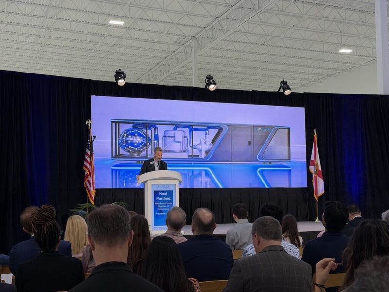 Noel Martinez, President of Palm Beach North Chamber of Commerce, speaks at the Ampera grand opening event, with a large product rendering displayed on screen behind him and an audience seated in the foreground